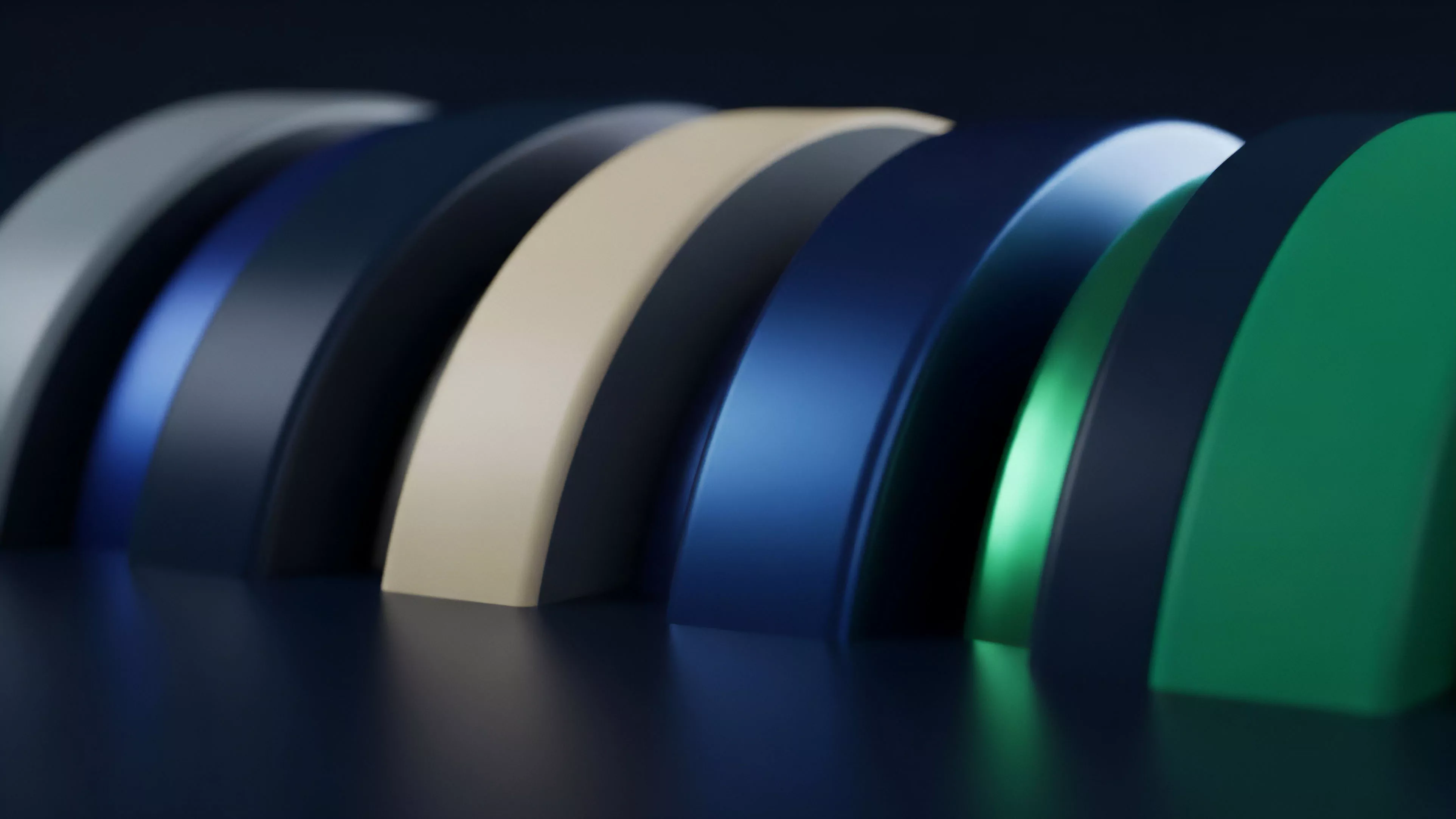 A row of layered, curved shapes in various colors, ranging from cool blues and greens to a warm beige, rests on a reflective dark surface. The shapes transition in color and texture, some appearing matte while others have a metallic sheen